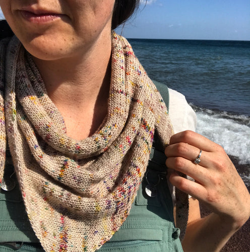 A close up shot of Artist's Point scarf around the neck of a freckled white woman with brown hair wearing sage-green overalls and a vintage diamond ring. Lake Superior pounds the North Shore in the background. 