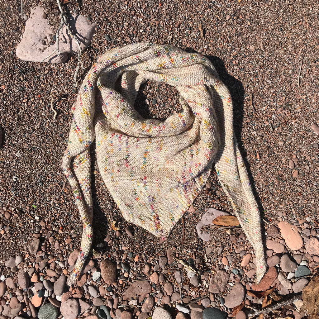 A loosly coiled knit triangle scarf rests in a transition zone of beach rock and pebbles. The hand-dyed yarn shows speckles of green, yellow, orange, purple, and pink against a warm neutral background. 