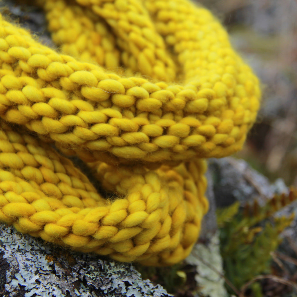 A doube-loop cowl knit in ochre yellow Malabrigo Rasta yarn drapes over a lichen speckled rock in Grand Marais, Minnesota. 